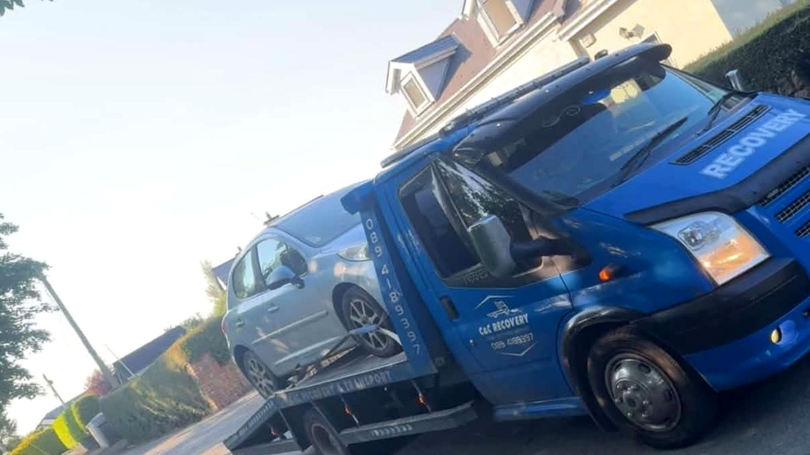 Blue C&C Recovery flatbed truck with a grey car loaded and strapped down on a road at Foulksmills, County Wexford
