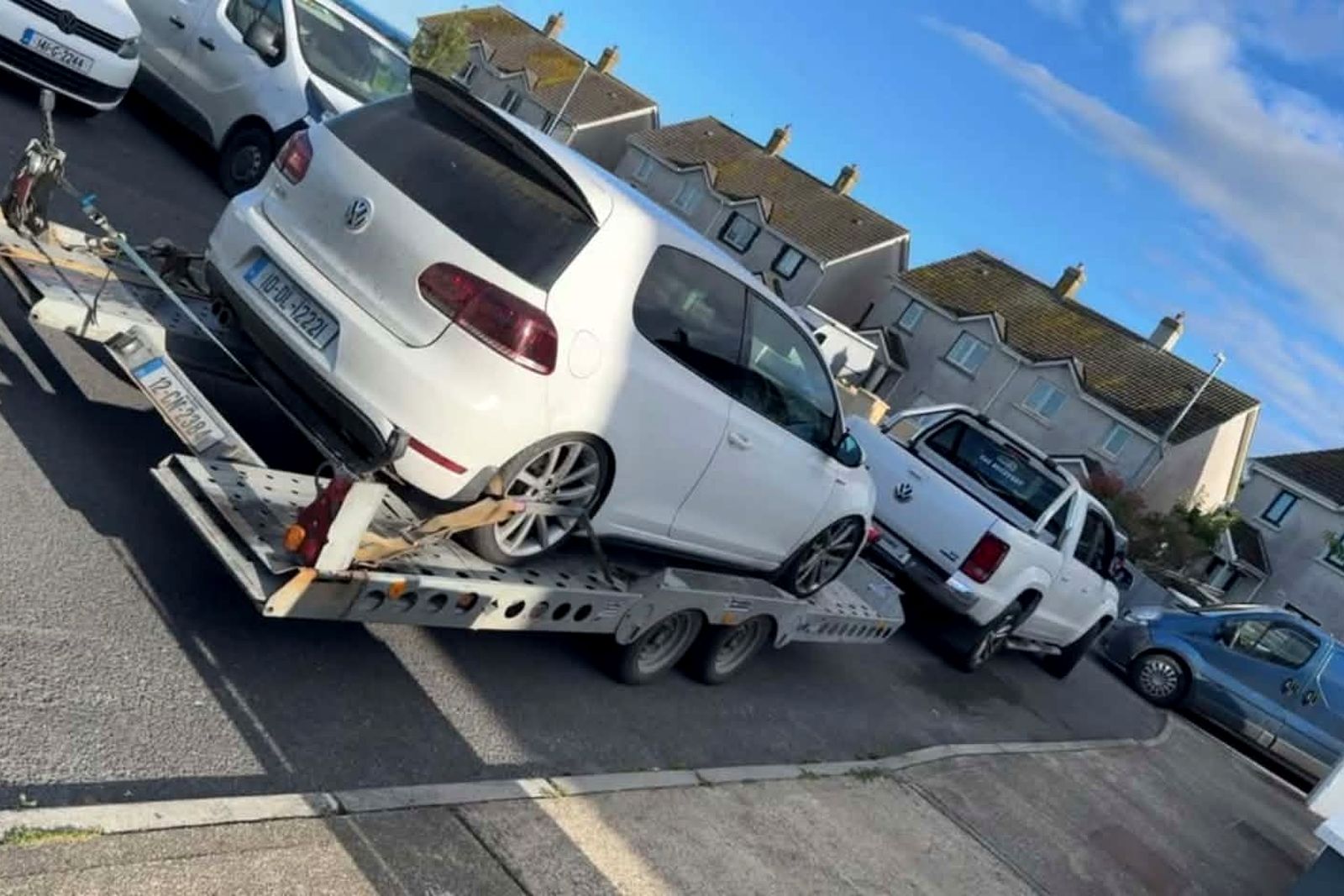 White Volkswagen Golf strapped down on a car transporter trailer behind a recovery pickup on a residential street